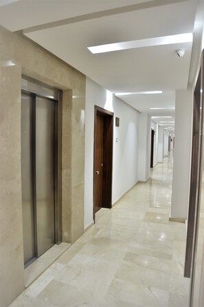 A long, well-lit corridor with a marble floor, an elevator, and a door on the right.