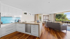 Eine moderne Küche mit weißen Schränken, einem eingebauten Geschirrspüler und einer großen Kochinsel mit Spüle. Ein Essbereich mit grauem Sofa und Blick auf das Meer durch große Fenster.