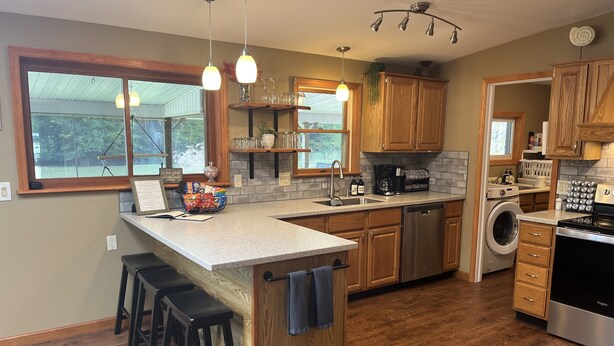 A modern kitchen with wooden cabinets, a built-in dishwasher, a washing machine, and a large island with bar stools. - Riverhouse NEW renovation, riverfront, game rm, fishing, swimming, paddle sports (Milford)