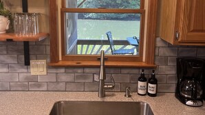 A kitchen with a stainless steel sink, wooden cabinets, a window with a view, and a hanging light fixture. - Riverhouse NEW renovation, riverfront, game rm, fishing, swimming, paddle sports (Milford)