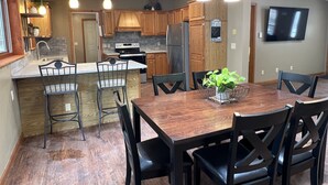 A modern kitchen with wooden cabinets, a central island, and a dining area with a wooden table and chairs. - Riverhouse NEW renovation, riverfront, game rm, fishing, swimming, paddle sports (Milford)