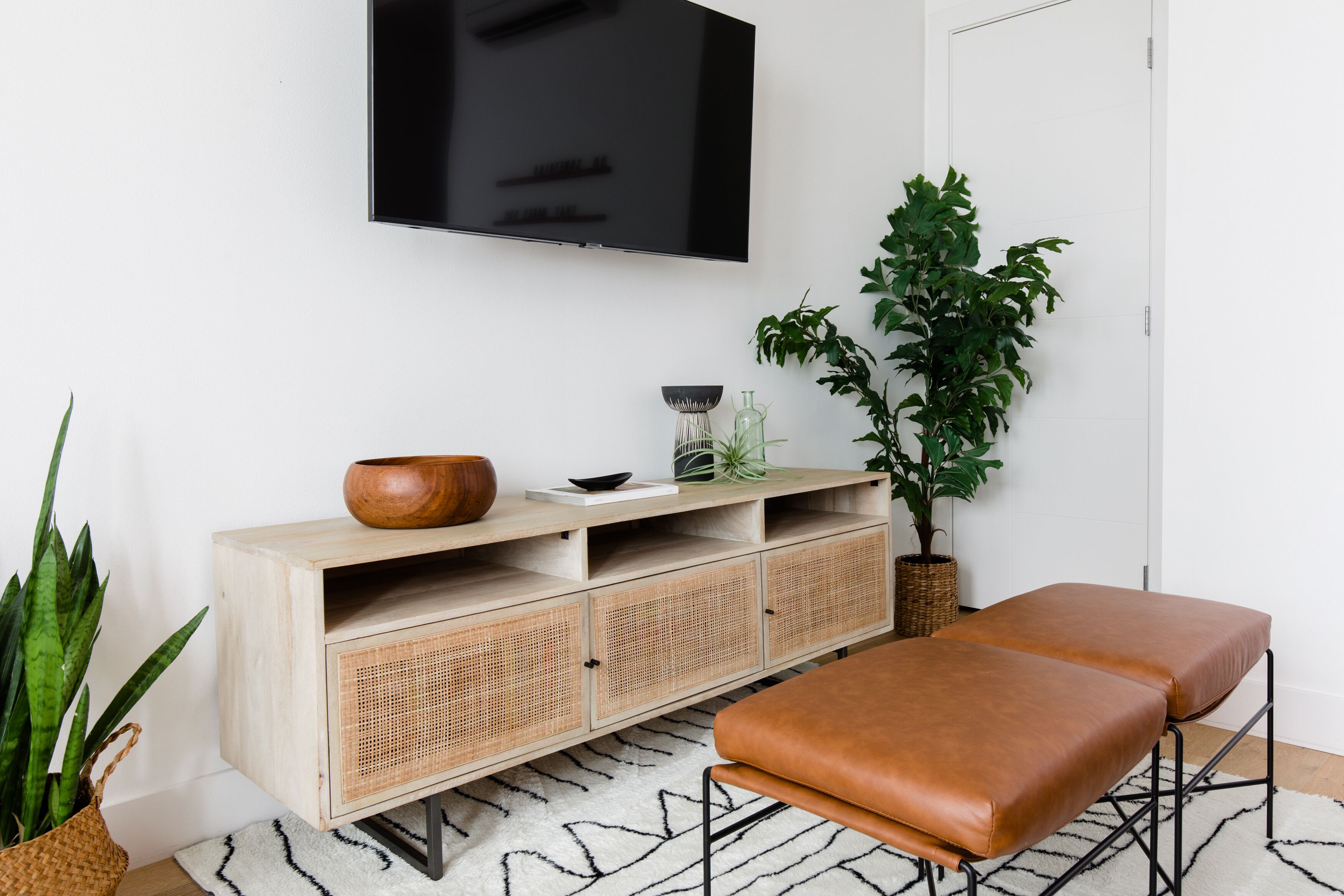 A modern living room with a wooden cabinet, a television, a brown leather bench, and potted plants.