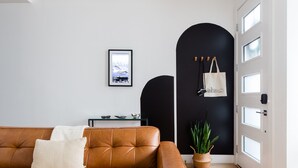 Un saló modern amb un sofà de cuir marró, una manta blanca i una taula auxiliar petita. Hi ha un quadre al mur, un arc negre i una porta blanca amb finestres.