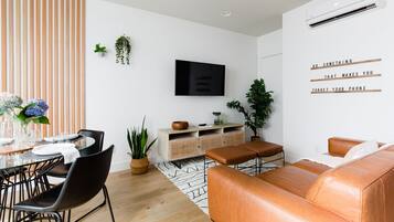 A modern living room with a TV, a sofa, a dining table with chairs, and a potted plant.
