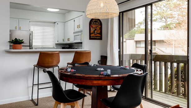 Een eethoek met een poker tafel, stoelen, een hanglamp en uitzicht op buiten door grote ramen.