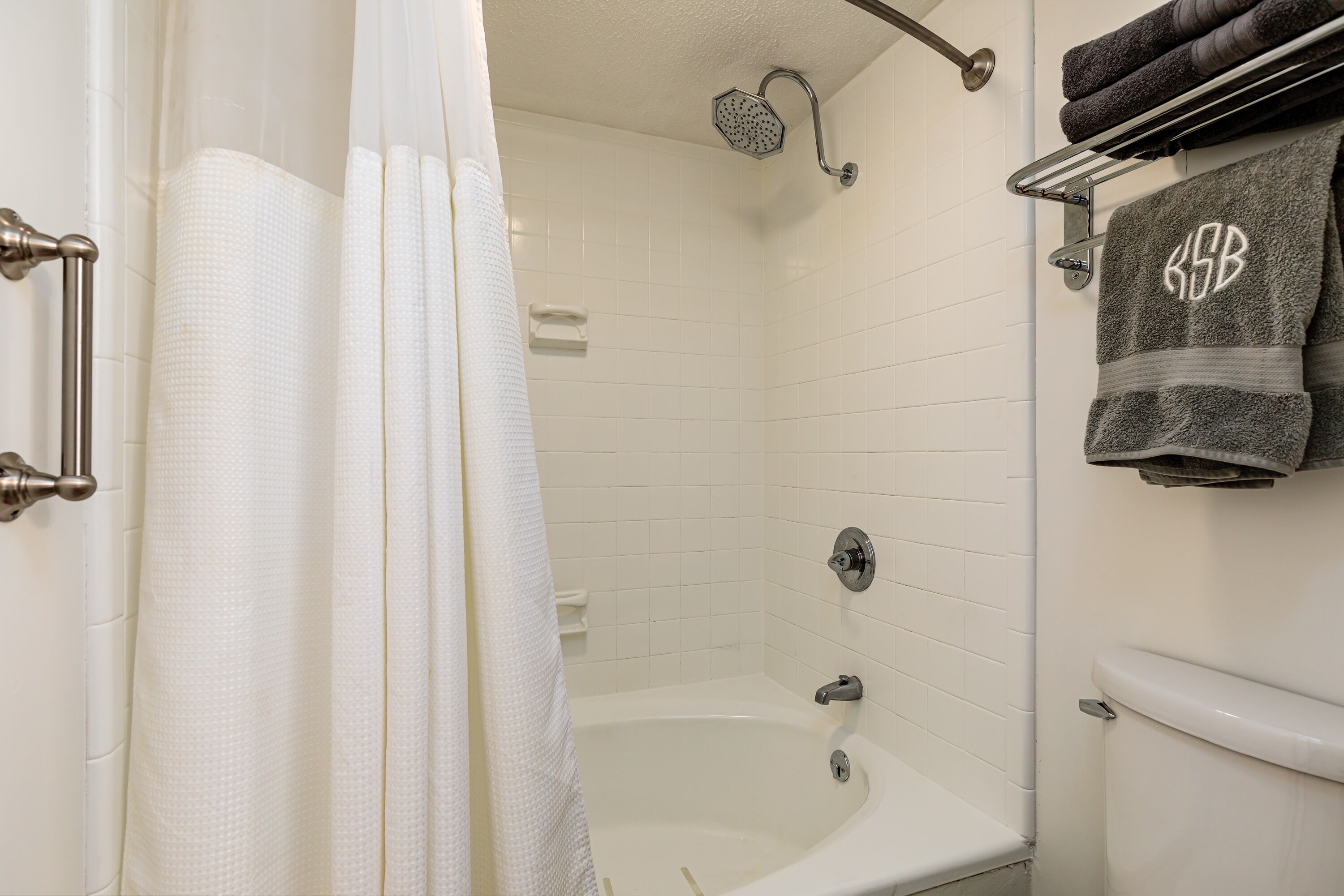 Ein Badezimmer mit Badewanne, Dusche und Toilette. Handtücher hängen an einem Wäscheständer, und ein Handtuchständer trägt ein Handtuch mit einem Monogramm.