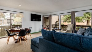 Een moderne woonkamer met een grote bank, een eettafel en stoelen. Er is een televisie aan de muur bevestigd en een balkon met tuinmeubilair.