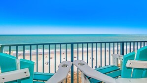 Een balkon met turquoise stoelen met uitzicht op een strand met mensen en de oceaan.