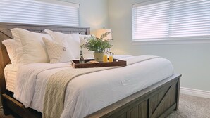 A bedroom with a wooden bed frame, white bedding, a bedside table with a lamp, and a window with blinds. - Double Down: Spacious Lakefront Retreat with Stunning Views & Hot Tub (Bella Vista)