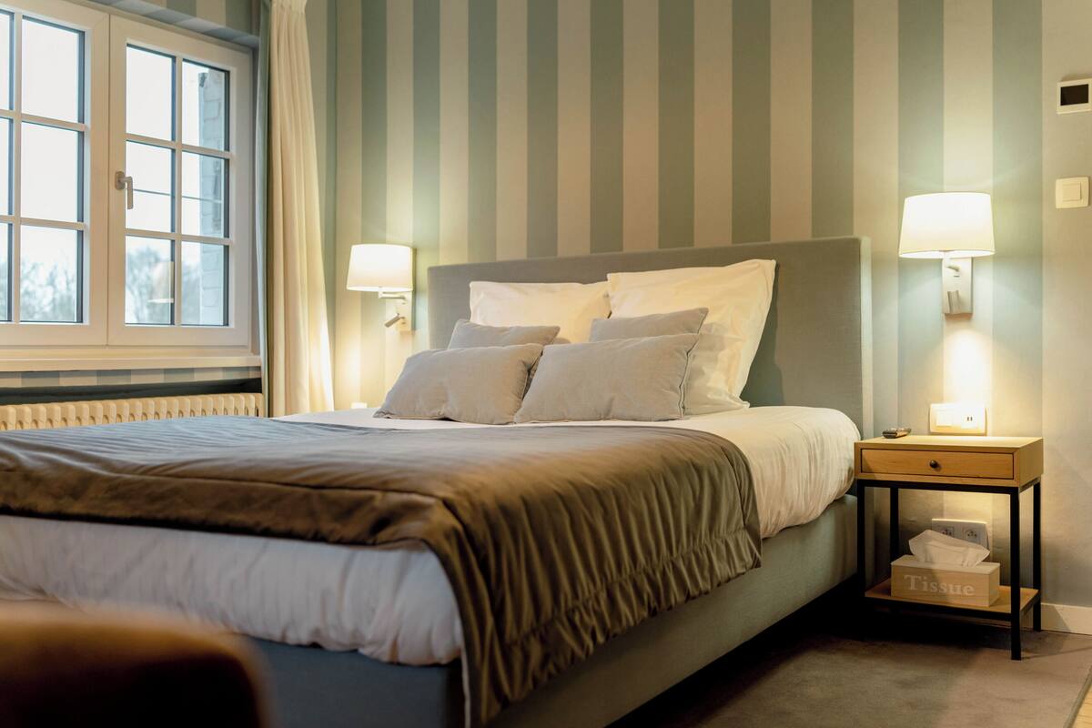 A neatly made bed with a headboard, two bedside lamps, a nightstand with a tissue box, and a window with curtains.