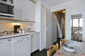 Eine kompakte Küche mit Mikrowelle, Kaffeemaschine und kleinem Herd. Ein Essbereich mit Tisch für zwei Personen, und eine Treppe, die in einen oberen Bereich führt.