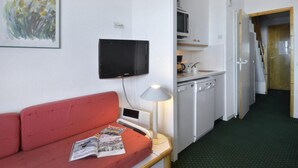 Quarto de hotel compacto com sofá vermelho, televisão de tela plana, uma mesinha com abajur, cozinha compacta e tapete verde.
