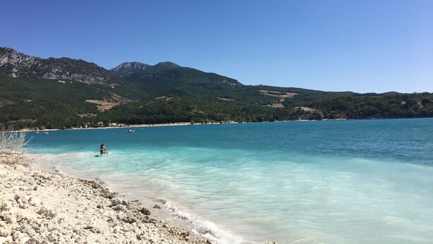 Een strand met helder turquoise water, een rotsachtige kustlijn en bergen op de achtergrond.