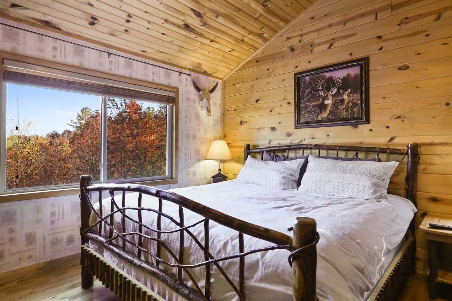 A wooden cabin bedroom with a bed, a window with a view of autumn trees, and a framed picture on the wall.