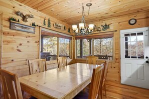 A wooden interior with a dining table, chairs, a chandelier, and windows with a view of trees. - Skyview: Full Cabin, Hot Tub, Game Room 5BR/5BA (Ellijay)