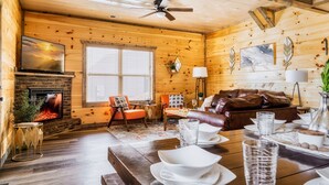 A cozy cabin interior with a fireplace, wooden walls, a ceiling fan, a dining area with a table set for four, and a leather sofa. - Paws Retreat: 5BR/3BA, Pvt Hot Tub - Sleeps 14 (sevierville)