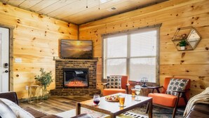 A cozy cabin interior with a stone fireplace, wooden walls, leather furniture, and a large window. - Paws Retreat: 5BR/3BA, Pvt Hot Tub - Sleeps 14 (sevierville)