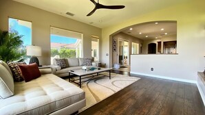 A spacious living room with a large sofa, a coffee table, and a ceiling fan. There is a view of the outside through large windows.