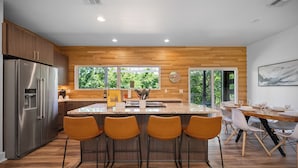 Una cocina moderna con una isla central, taburetes de bar, electrodomésticos de acero inoxidable y grandes ventanales que ofrecen vistas a zonas verdes.