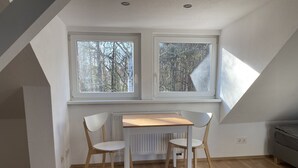 A small, bright room with a sloped ceiling, two windows with a view of trees, a wooden table with two chairs, and a radiator under the windows. - Zeuthen, top location, high-quality/quiet/bright furnished top floor apartment with garden, near the lake (Zeuthen)