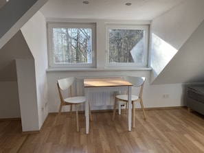 Una habitación pequeña y luminosa con techo inclinado, dos ventanas que dan a árboles, una mesa de madera con dos sillas y un radiador debajo de las ventanas.