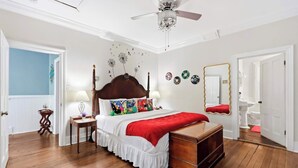 A bedroom with a wooden bed, a dresser, a ceiling fan, a mirror, and a door leading to a bathroom. - The House of Hue | 1870s Artful Savannah Retreat (Savannah)