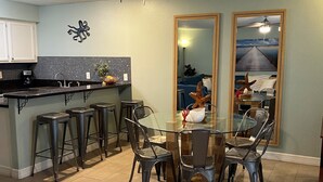 A dining area with a glass table and metal chairs, a ceiling fan, and a kitchen with white cabinets and a black countertop. - Cognito Townhouse 3 Bedroom Home by RedAwning (South Padre Island)