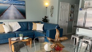 A living room with a blue sofa, a glass dining table, and a kitchen area with a refrigerator. - Cognito Townhouse 3 Bedroom Home by RedAwning (South Padre Island)