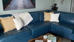 A living room with a blue leather sofa, a wooden coffee table, and a painting of a pier. - Cognito Townhouse 3 Bedroom Home by RedAwning (South Padre Island)