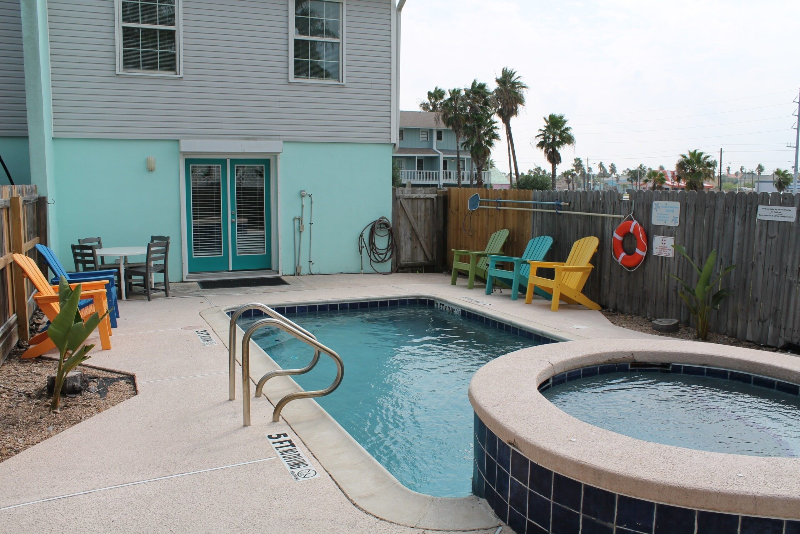 A swimming pool with a hot tub, surrounded by a wooden fence, and a house with a teal exterior.