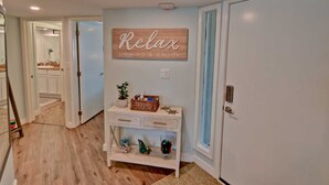 A hallway with a wooden sign that reads "Relax," a small white console table with decorative items, and a door with a light switch. - Gulfpoint 3209 2 Bedroom Condo by RedAwning (South Padre Island)