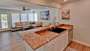 A modern kitchen with granite countertops, a built-in sink, and white cabinetry. There is a dining area with a sofa and a ceiling fan. - Gulfpoint 3209 2 Bedroom Condo by RedAwning (South Padre Island)