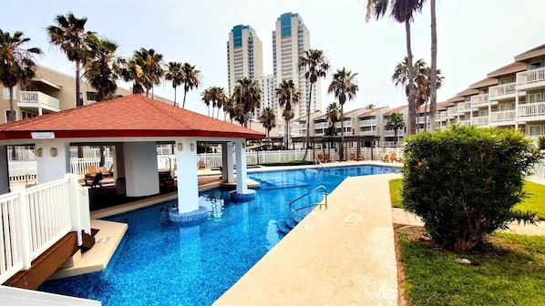 Una piscina con un pabellón de techo rojo, rodeada de palmeras y edificios de apartamentos modernos.