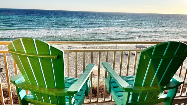 Twee groene Adirondack-stoelen op een houten terras met uitzicht op een strand en golven.