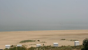 A sandy beach with several white structures, likely beach huts, and a distant sailboat on the water. - Lovely Beach House for 4 persons within walking distance from the Beach  (Breskens)