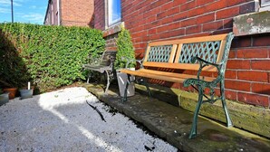 Eine Bank vor einem Backsteinhaus, mit einer Hecke und einigen Kübelpflanzen in der Nähe.
