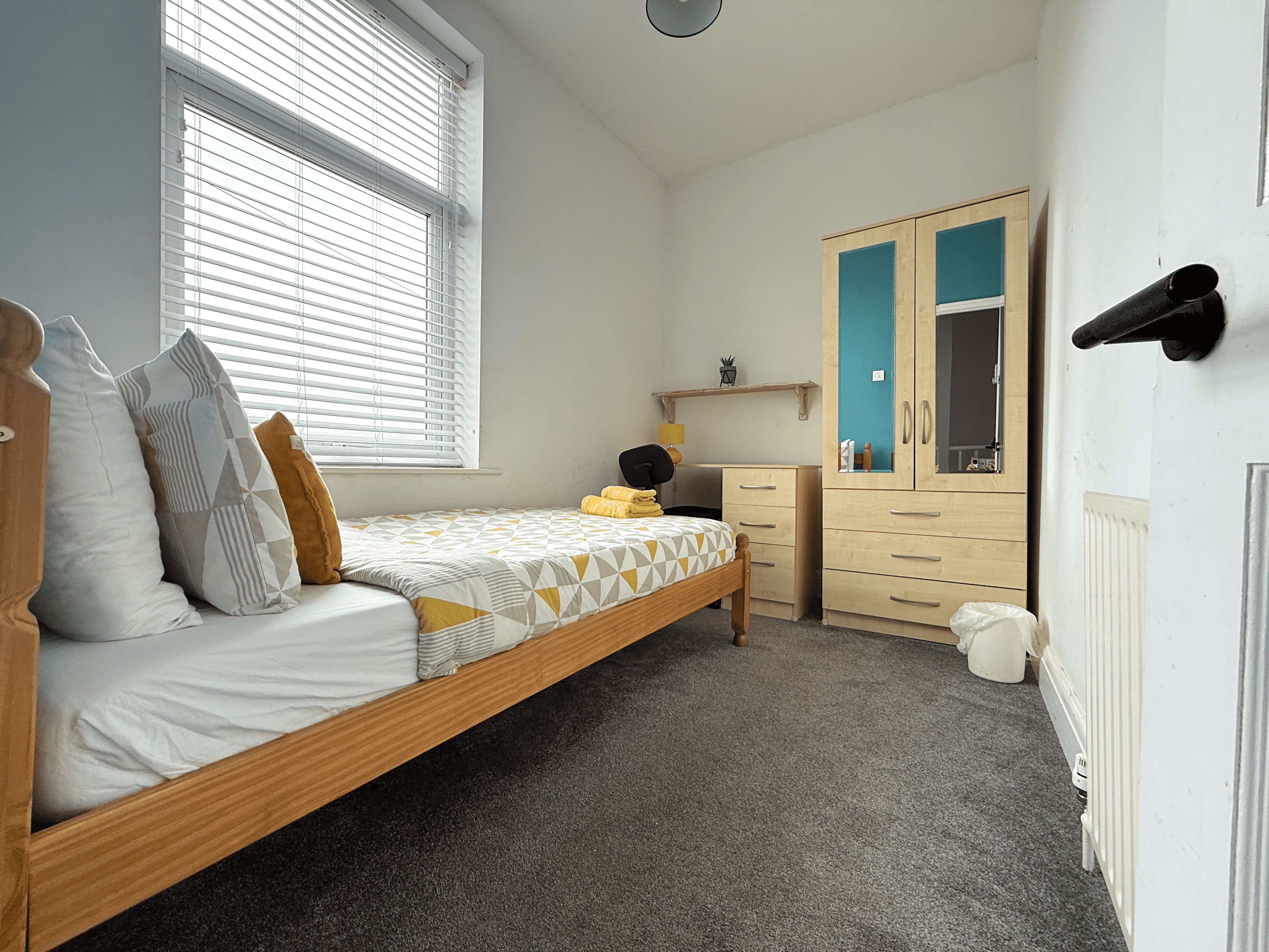 A bedroom with a single bed, a dresser, a wardrobe, and a window with blinds.