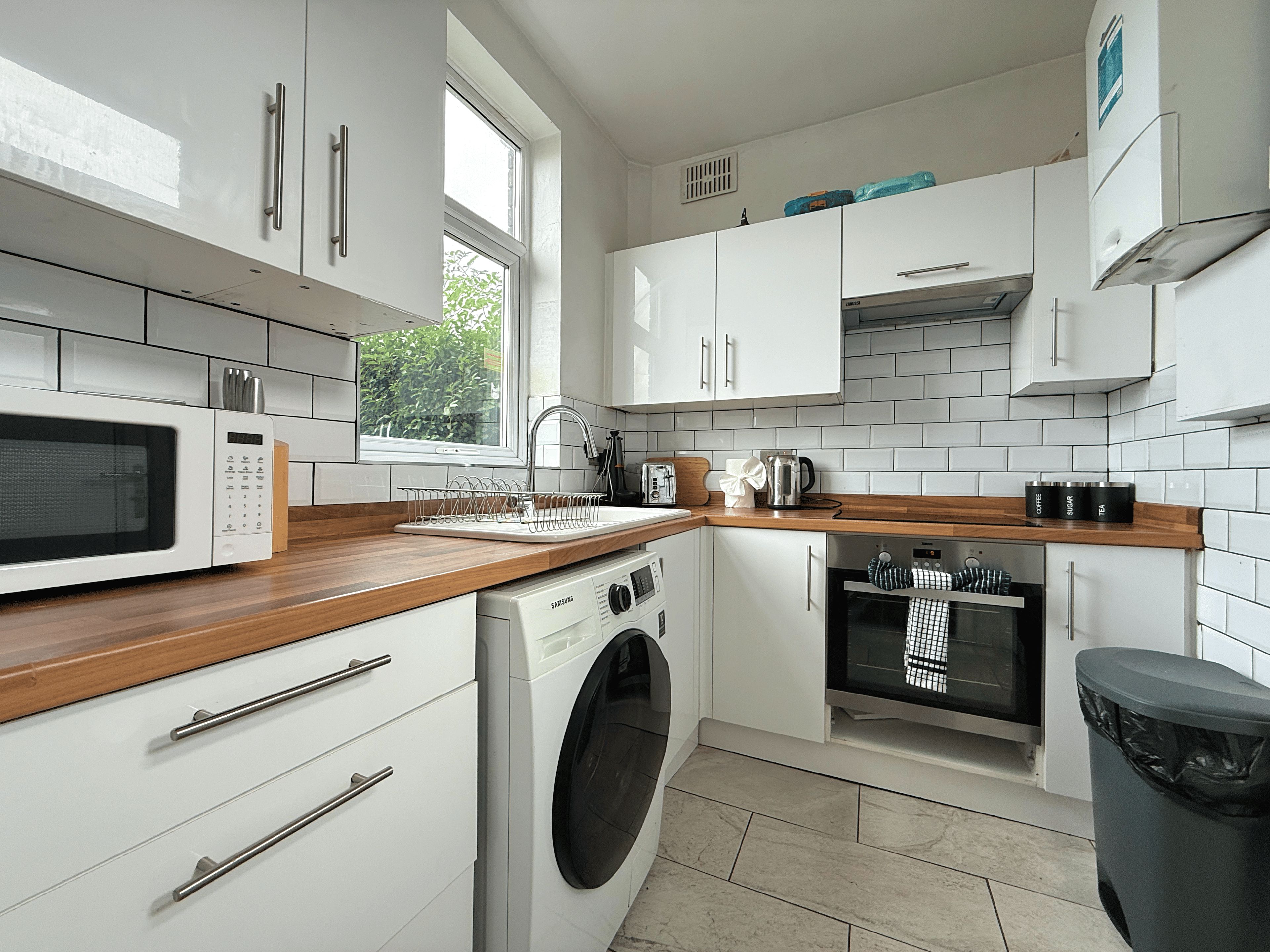 A modern kitchen with white cabinetry, a built-in microwave, a washing machine, and a window providing natural light.