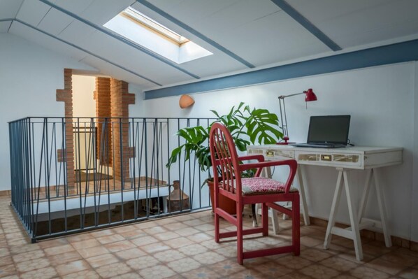 Una habitación con tragaluz, un escritorio con una computadora portátil, una silla roja y una planta en maceta.