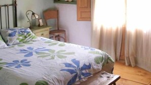 Una camera da letto con un letto, una panca di legno, una sedia, un comodino, una lampada e una finestra con tende.