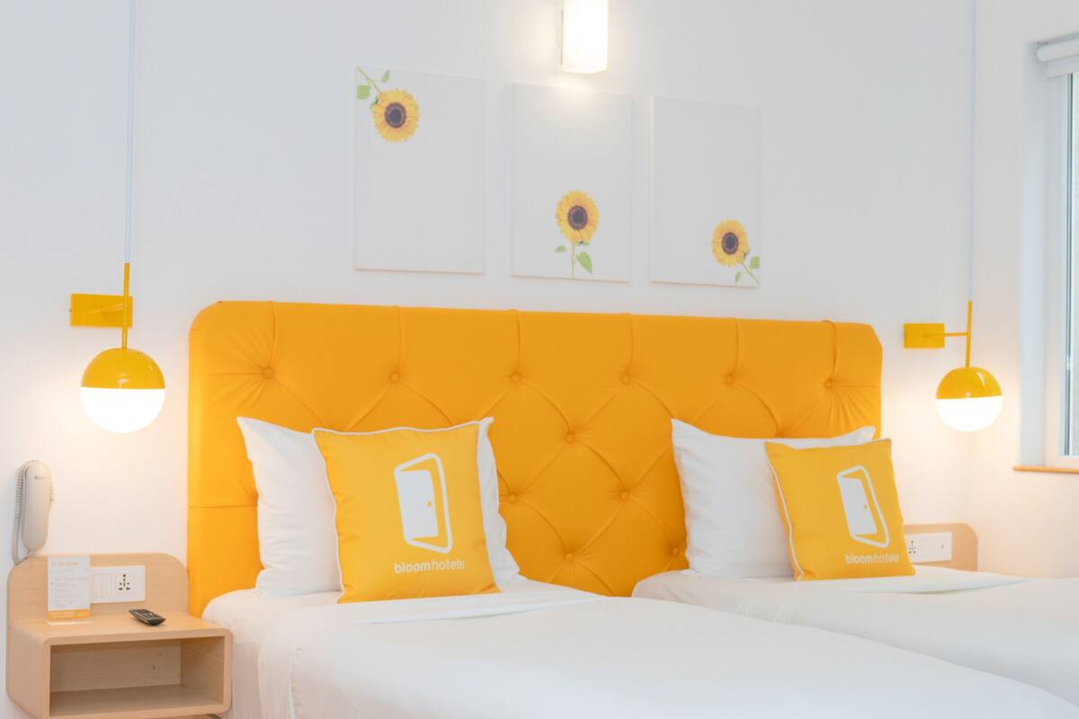 Quarto de hotel com cabeceira laranja vibrante, roupa de cama branca e arte de parede com girassóis.