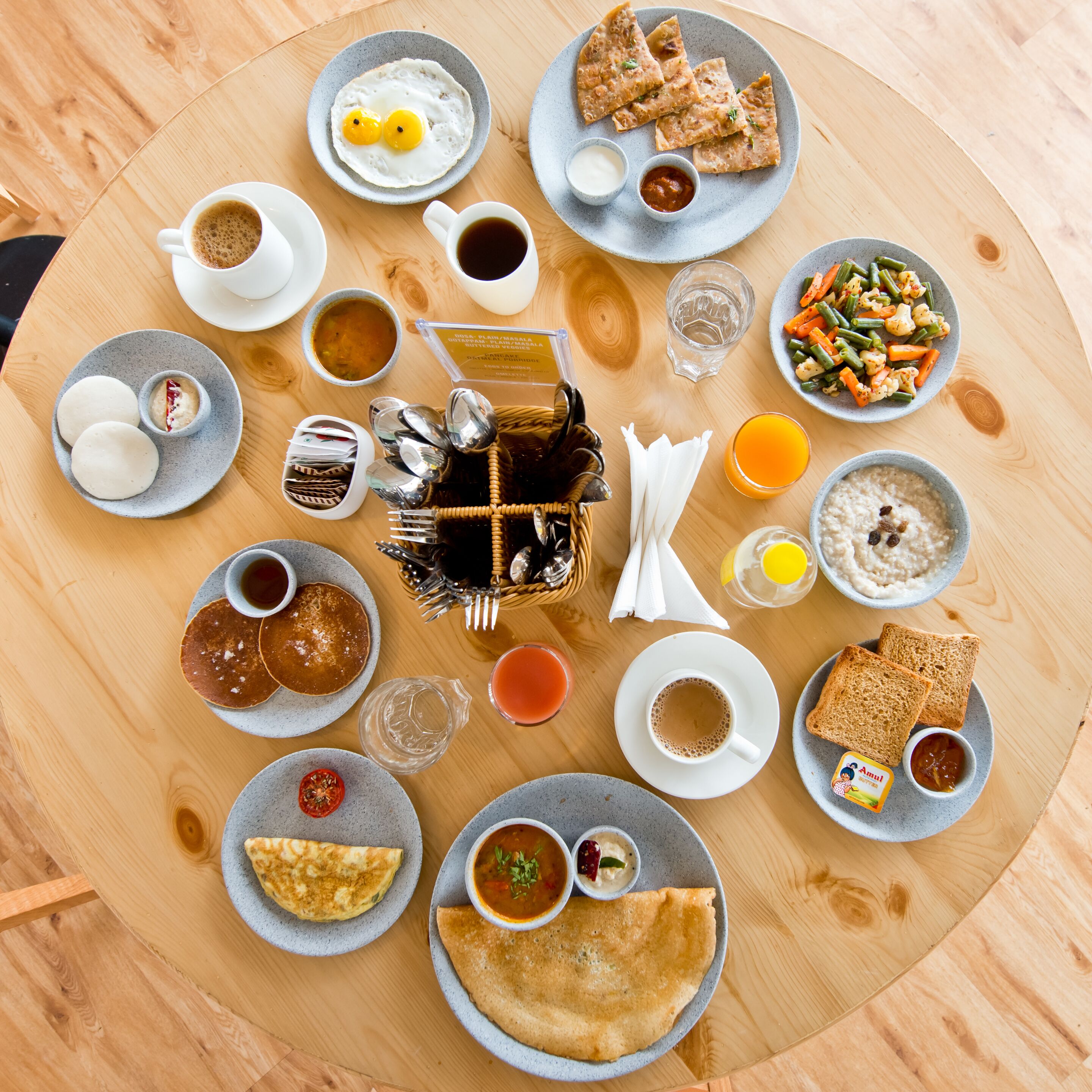 A breakfast spread on a round wooden table with various dishes including pancakes, omelettes, toast, and a variety of sides and beverages.