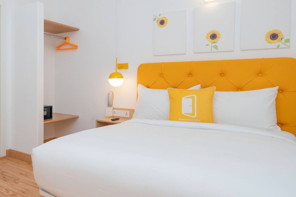 Quarto de hotel com uma cama grande, uma mesinha de cabeceira com abajur, um lustre e três quadros de girassóis na parede.