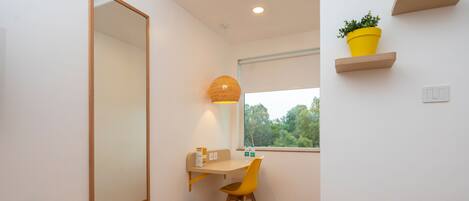A modern, minimalistic room with a wooden floor, a desk with a chair, a large mirror, an air conditioning unit, a window with a view of trees, and yellow plant pots on shelves.
