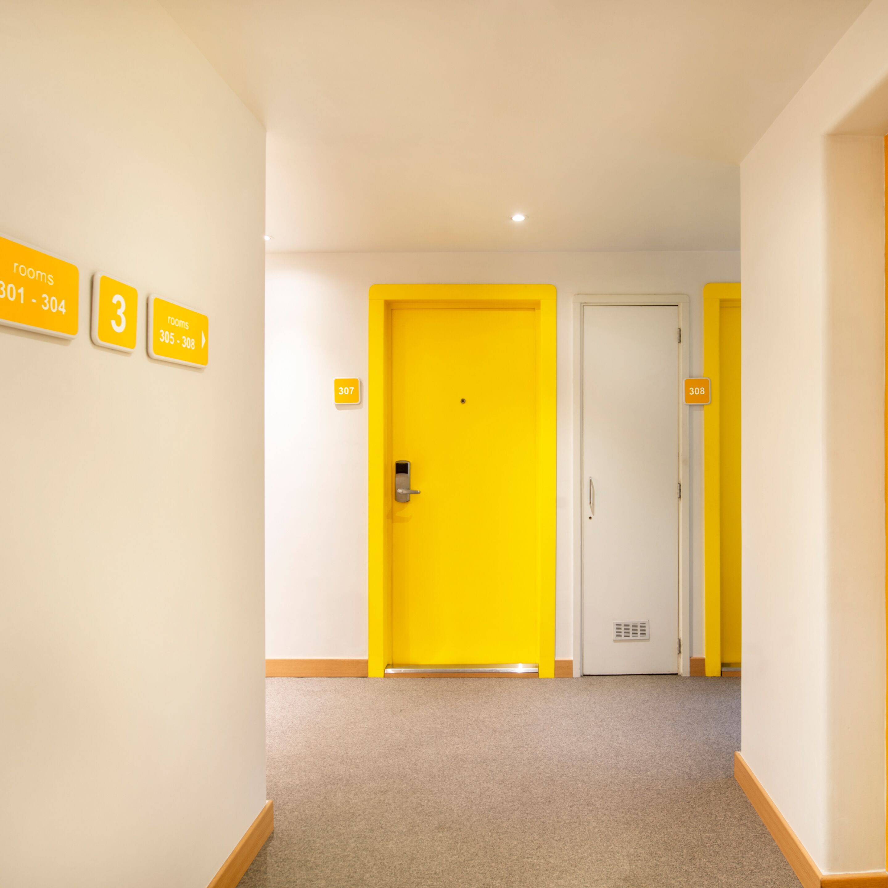 Ein Flur mit einer leuchtend gelben Tür mit der Beschriftung 307, einer weißen Tür mit der Beschriftung 308 und einem Schild, das die Zimmer 301 bis 304 und 305 bis 308 ausweist.