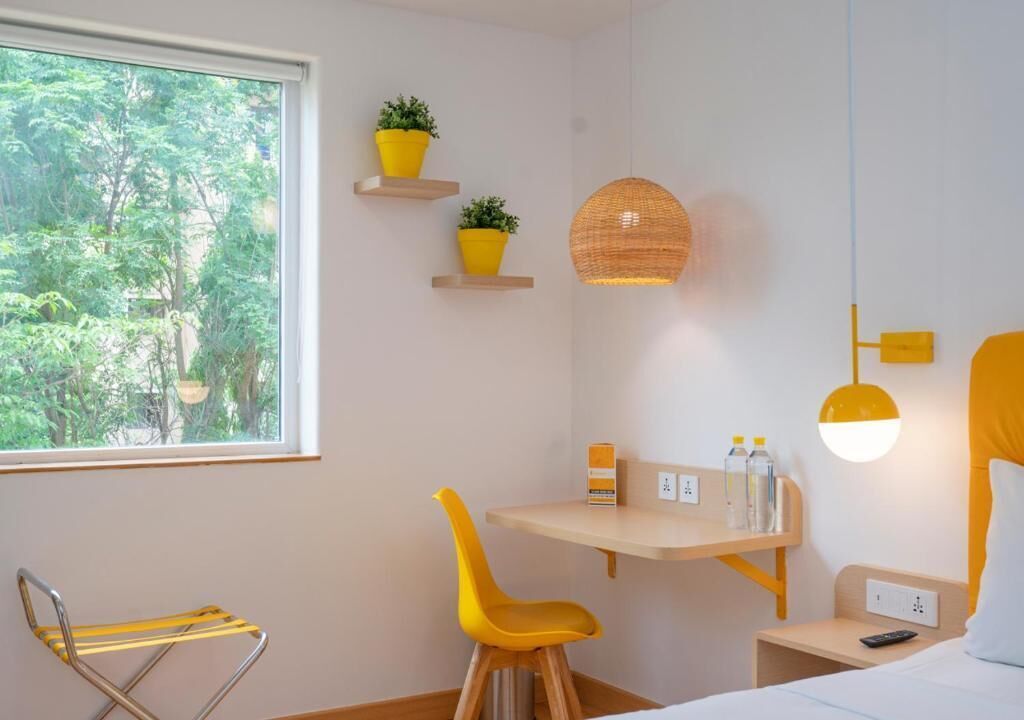 A room with a bed, a desk with a chair, a hanging lamp, and a window with a view of trees.