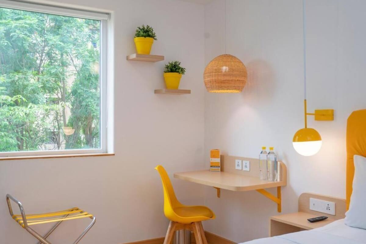 Quarto com cama, escrivaninha com cadeira, luminária pendente e janela com vista para árvores.