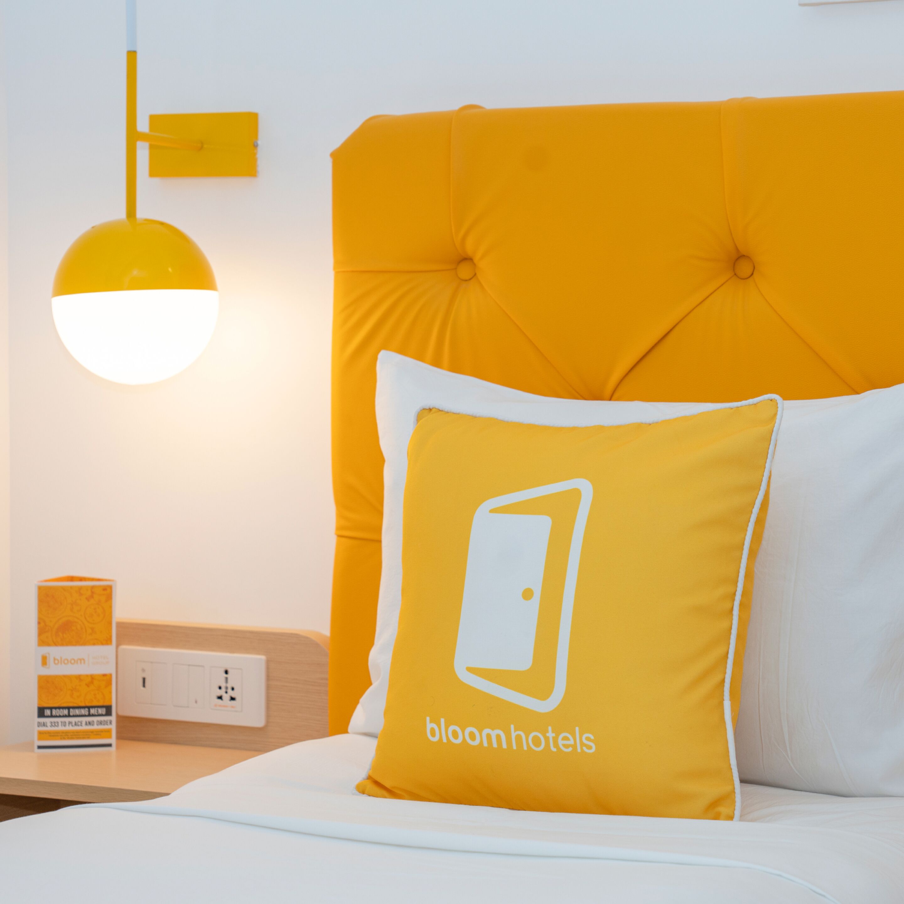 Una habitación de hotel con cabecera capitonada, una mesita de noche con lámpara y una almohada con logotipo.