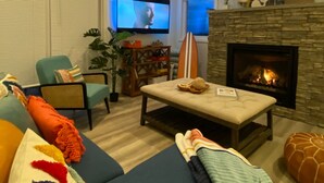 A living room with a stone fireplace, a television mounted on the wall, and a large window with blinds.