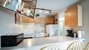 A modern kitchen with wooden cabinets, a white countertop, a microwave, a stove, a refrigerator, and a sink. There are two wooden chairs at the table.